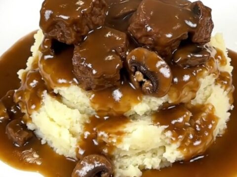 A close-up of tender beef tips and mushrooms covered in a rich brown gravy served over creamy mashed potatoes.