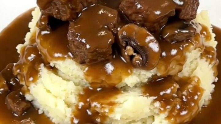 A close-up of tender beef tips and mushrooms covered in a rich brown gravy served over creamy mashed potatoes.