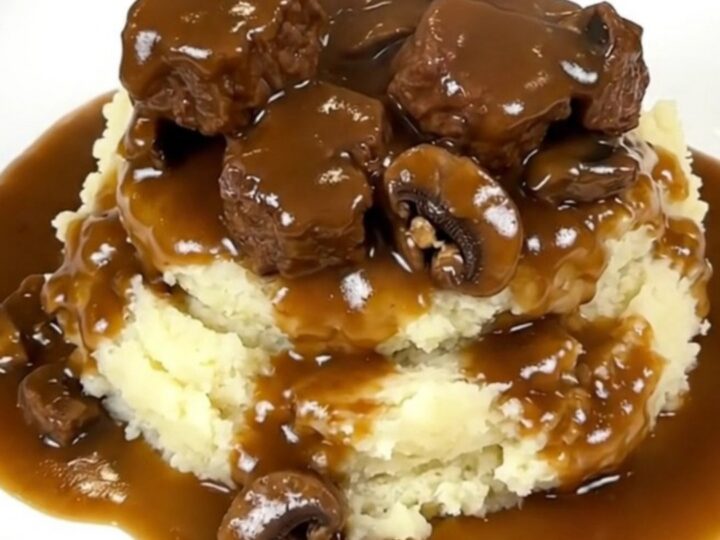 A close-up of tender beef tips and mushrooms covered in a rich brown gravy served over creamy mashed potatoes.