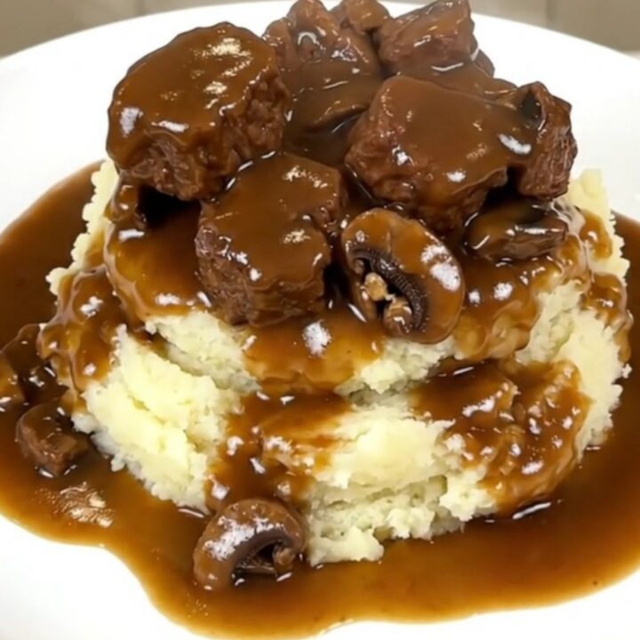 A close-up of tender beef tips and mushrooms covered in a rich brown gravy served over creamy mashed potatoes.