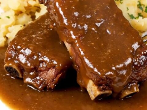 A close-up of tender country-style pork ribs and baby potatoes covered in a rich brown gravy on a white plate.