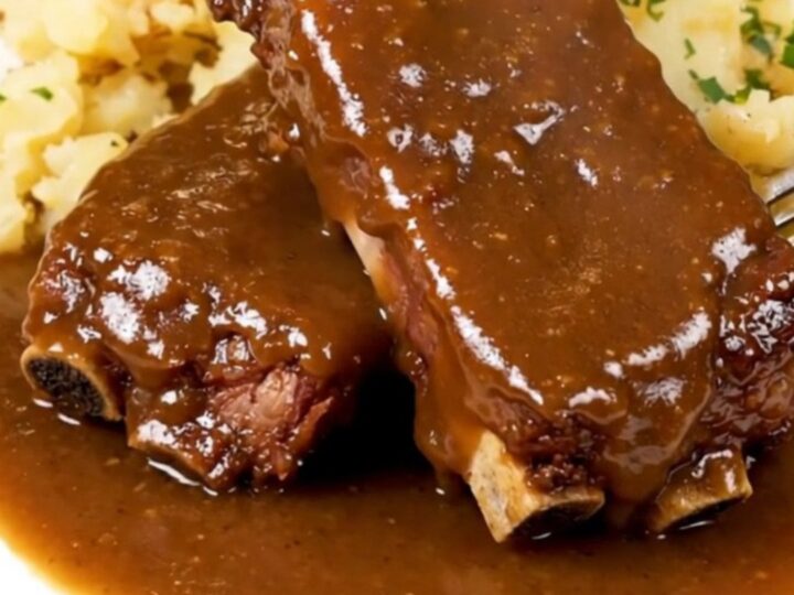 A close-up of tender country-style pork ribs and baby potatoes covered in a rich brown gravy on a white plate.