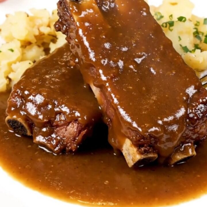 A close-up of tender country-style pork ribs and baby potatoes covered in a rich brown gravy on a white plate.