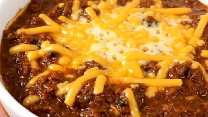 A bowl of hearty baked potato beef chili topped with melted cheddar cheese on a wooden table.