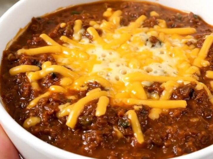 A bowl of hearty baked potato beef chili topped with melted cheddar cheese on a wooden table.