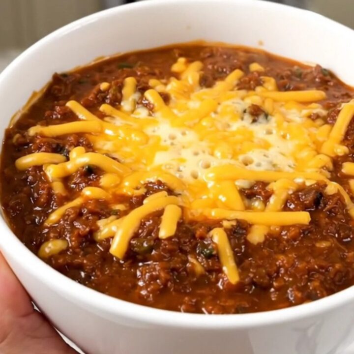 A bowl of hearty baked potato beef chili topped with melted cheddar cheese on a wooden table.