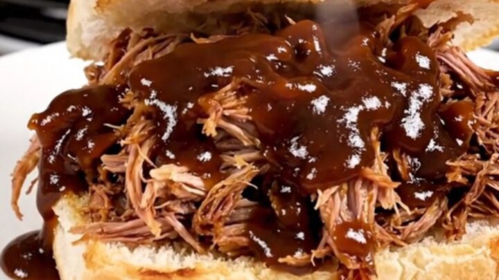A close-up of glossy, steaming shredded pulled pork piled high on a soft potato roll on a white plate.