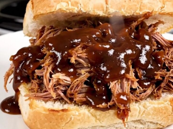 A close-up of glossy, steaming shredded pulled pork piled high on a soft potato roll on a white plate.
