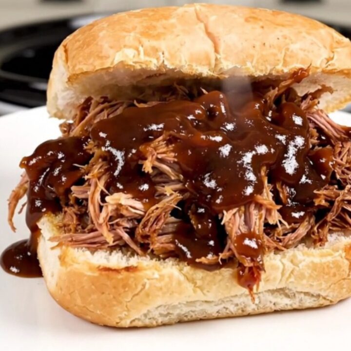 A close-up of glossy, steaming shredded pulled pork piled high on a soft potato roll on a white plate.