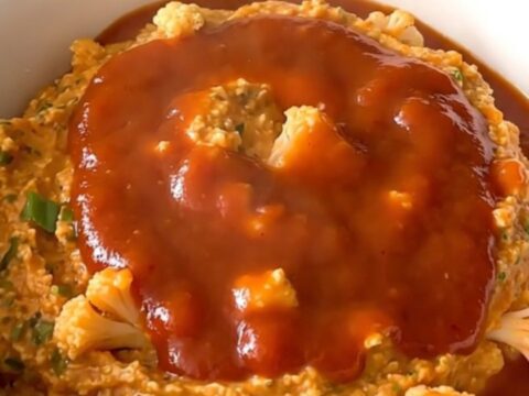 A top-down view of a black slow cooker filled with raw cauliflower, green onions, and buffalo sauce.
