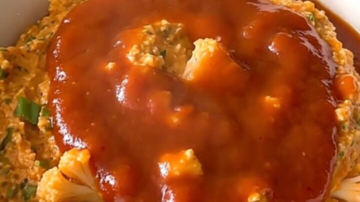 A top-down view of a black slow cooker filled with raw cauliflower, green onions, and buffalo sauce.