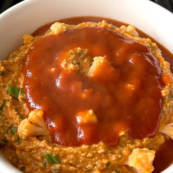 A top-down view of a black slow cooker filled with raw cauliflower, green onions, and buffalo sauce.