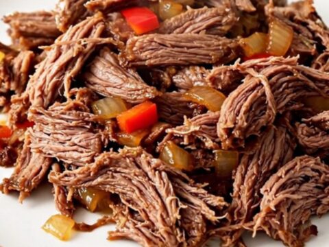 A top-down view of a black slow cooker filled with raw flank steak, sliced peppers, onions, and salsa.