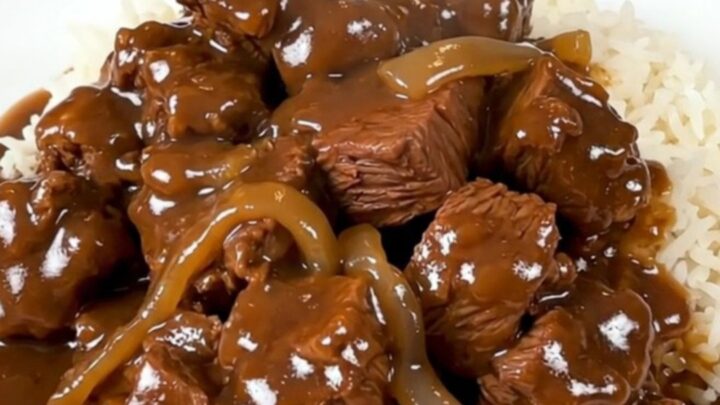A slow cooker filled with tender beef cubes, thick sweet onion slices, and a rich, savory brown gravy.