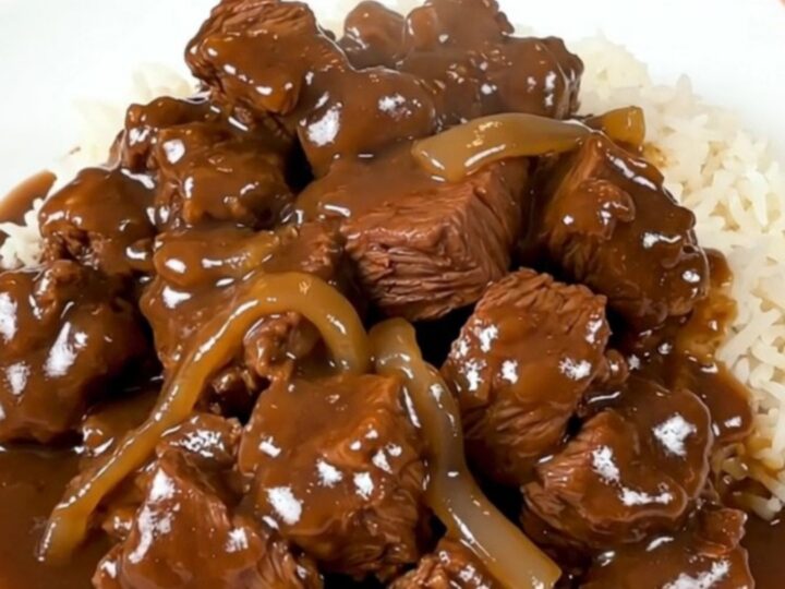 A slow cooker filled with tender beef cubes, thick sweet onion slices, and a rich, savory brown gravy.