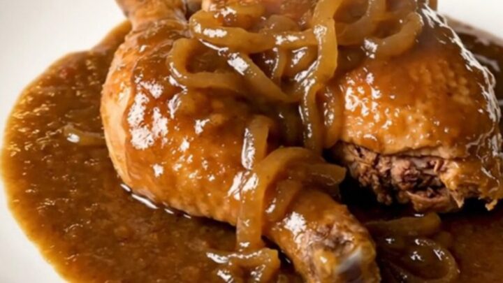 Slow cooked chicken thighs covered in a rich brown onion gravy in a black slow cooker on a white countertop.