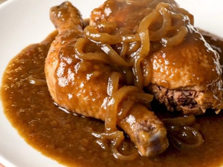 Slow cooked chicken thighs covered in a rich brown onion gravy in a black slow cooker on a white countertop.
