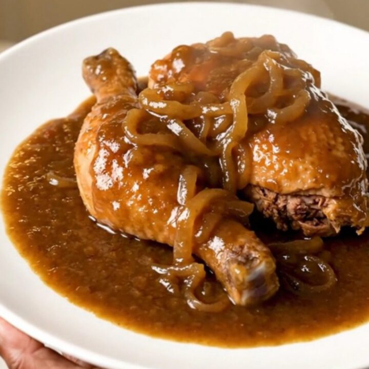Slow cooked chicken thighs covered in a rich brown onion gravy in a black slow cooker on a white countertop.