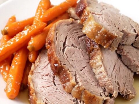A close-up of a perfectly cooked, juicy garlic herb pork roast sitting on a white plate with glazed carrots.