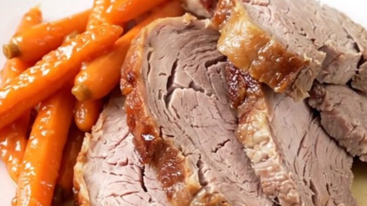 A close-up of a perfectly cooked, juicy garlic herb pork roast sitting on a white plate with glazed carrots.