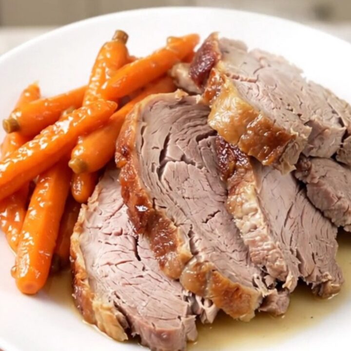 A close-up of a perfectly cooked, juicy garlic herb pork roast sitting on a white plate with glazed carrots.
