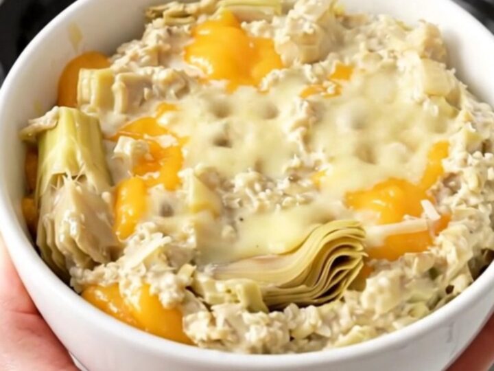 A top-down view of a black slow cooker filled with a creamy artichoke dip on a speckled countertop.