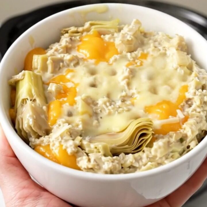 A top-down view of a black slow cooker filled with a creamy artichoke dip on a speckled countertop.