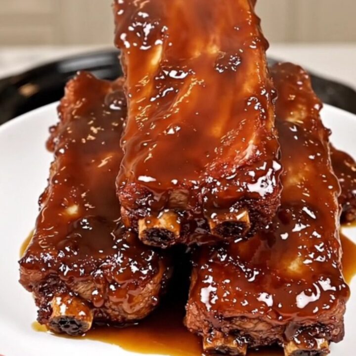 A top-down view of raw baby back ribs being seasoned in a black slow cooker on a white quartz countertop.