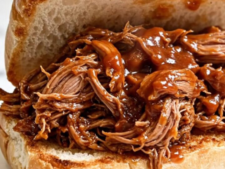 A close-up view of succulent, shredded pulled pork piled high on a toasted roll, showing a glossy barbecue sauce finish.