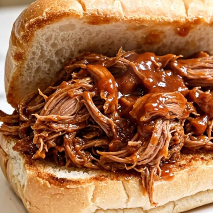 A close-up view of succulent, shredded pulled pork piled high on a toasted roll, showing a glossy barbecue sauce finish.