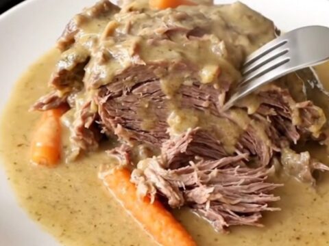 A close-up of a tender beef chuck roast covered in creamy gravy with baby carrots in a slow cooker.