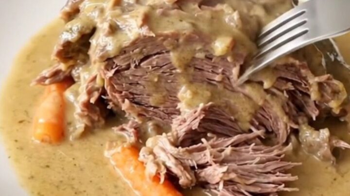 A close-up of a tender beef chuck roast covered in creamy gravy with baby carrots in a slow cooker.