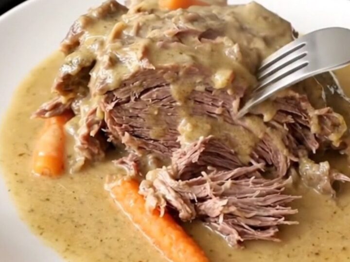A close-up of a tender beef chuck roast covered in creamy gravy with baby carrots in a slow cooker.