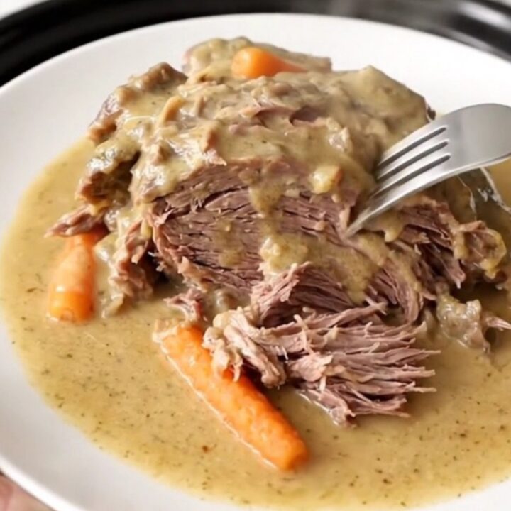 A close-up of a tender beef chuck roast covered in creamy gravy with baby carrots in a slow cooker.