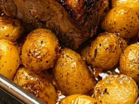 Perfectly roasted pork loin surrounded by glistening, blistered baby potatoes in a dark roasting pan