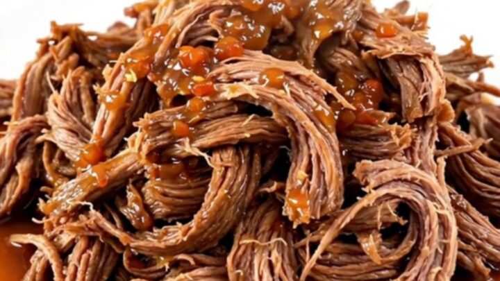 A close-up of tender, shredded sweet chili beef piled on a white plate with a soft-focus kitchen background.