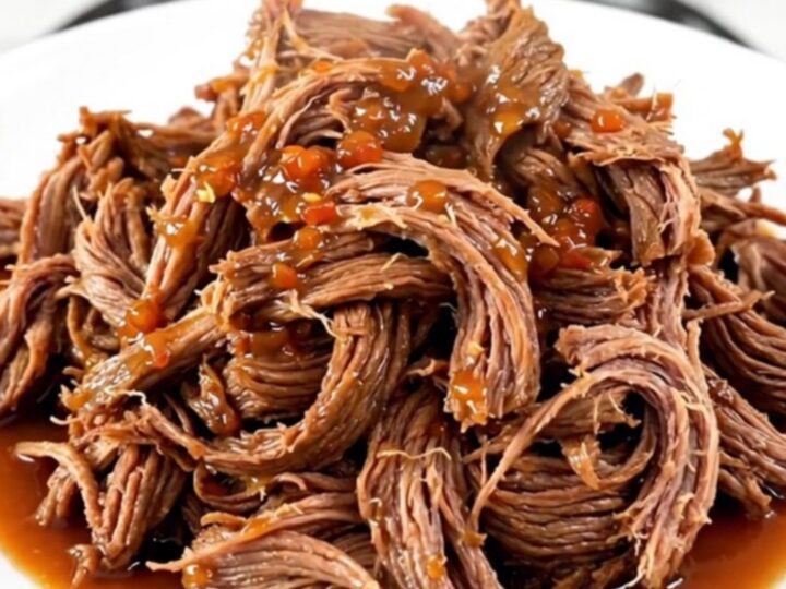 A close-up of tender, shredded sweet chili beef piled on a white plate with a soft-focus kitchen background.