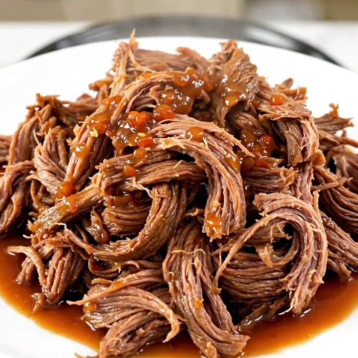 A close-up of tender, shredded sweet chili beef piled on a white plate with a soft-focus kitchen background.