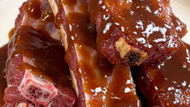 A plate of tender, glazed baby back ribs resting on a white plate against a blurred kitchen background.
