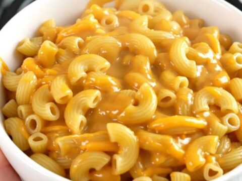 A top-down view of a black slow cooker filled with raw macaroni, shredded cheddar, and beer, ready to be cooked.
