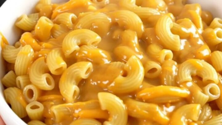 A top-down view of a black slow cooker filled with raw macaroni, shredded cheddar, and beer, ready to be cooked.