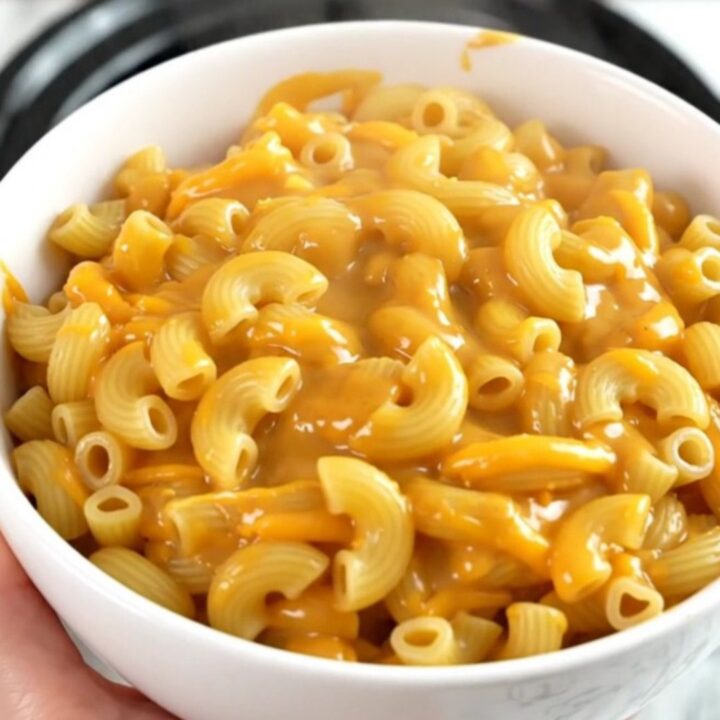 A top-down view of a black slow cooker filled with raw macaroni, shredded cheddar, and beer, ready to be cooked.