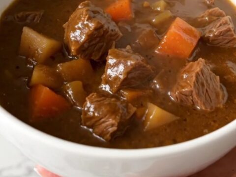 A steaming, hearty beef stew with chunks of tender beef, baby potatoes, and bright carrots in a deep white bowl.