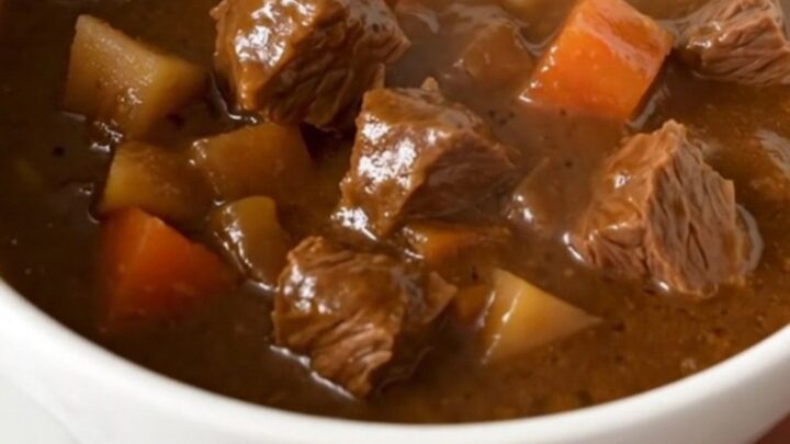 A steaming, hearty beef stew with chunks of tender beef, baby potatoes, and bright carrots in a deep white bowl.