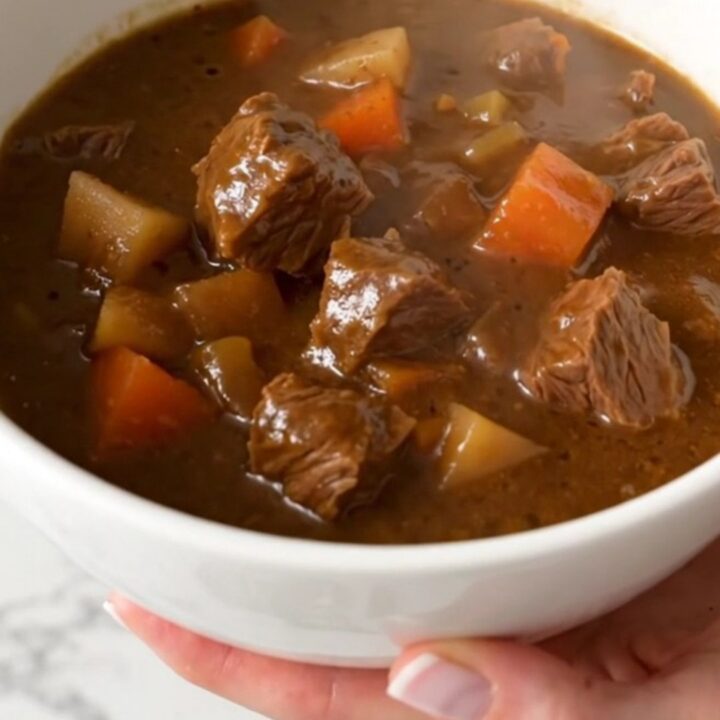 A steaming, hearty beef stew with chunks of tender beef, baby potatoes, and bright carrots in a deep white bowl.