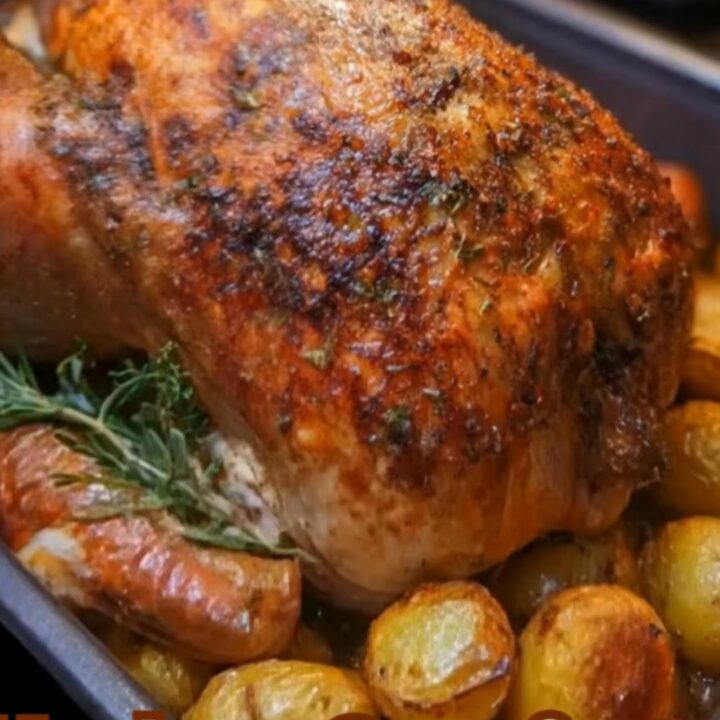 A whole golden-brown roast chicken in a dark baking pan surrounded by glistening baby potatoes and fresh rosemary.