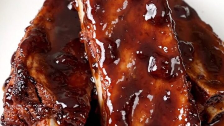 Slow cooker ribs glazed in a shiny, dark cherry cola sauce on a white plate.
