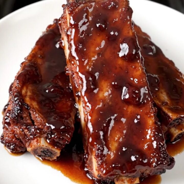 Slow cooker ribs glazed in a shiny, dark cherry cola sauce on a white plate.