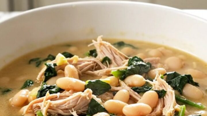A white bowl filled with steaming Tuscan chicken and white bean stew with visible pieces of shredded chicken and spinach.
