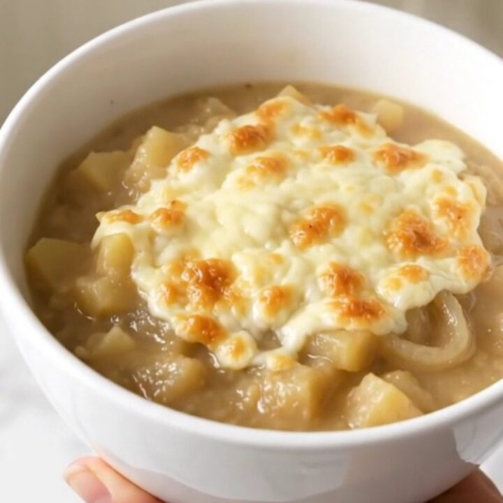 A steaming bowl of creamy French onion potato chowder topped with melted cheese on a white background.
